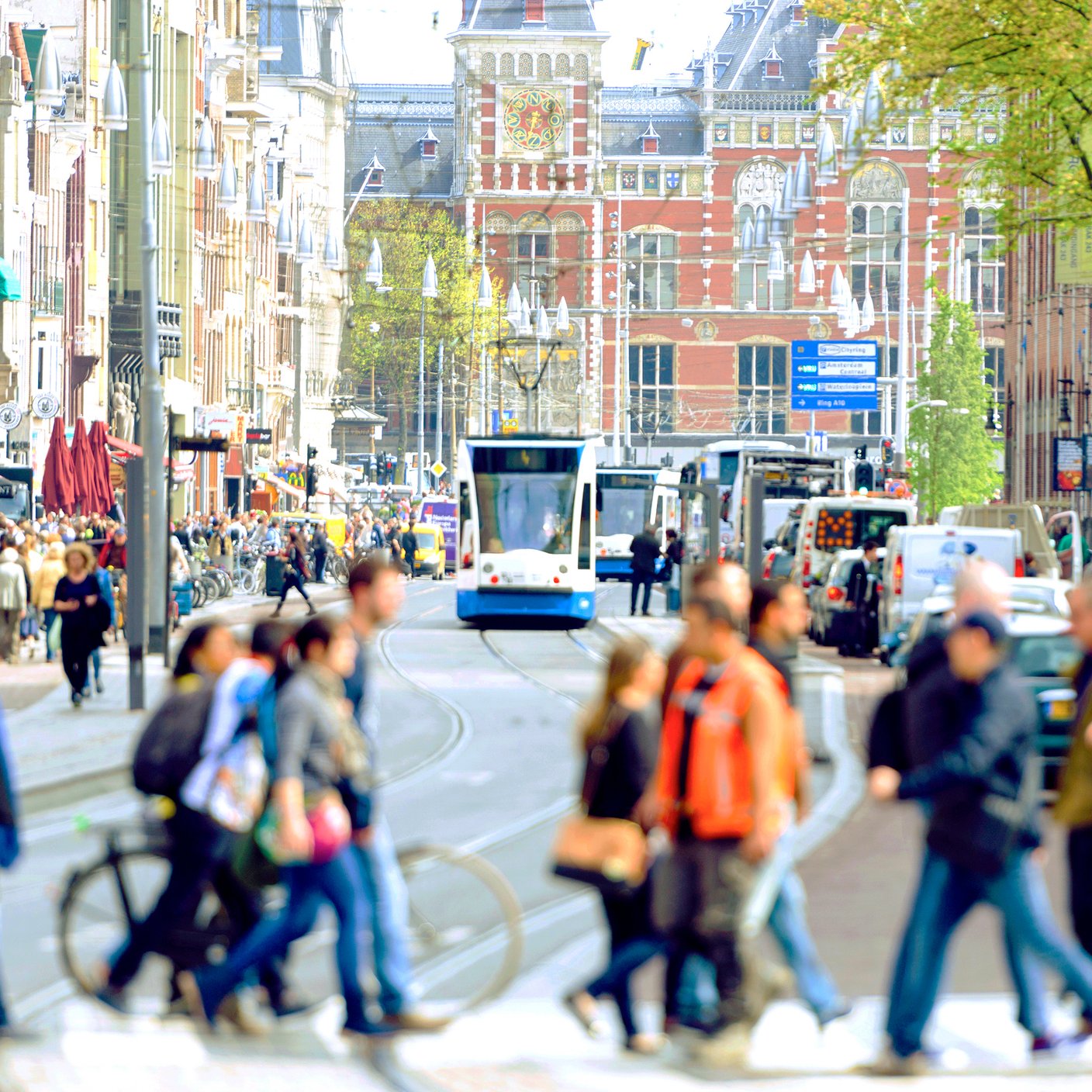 The famous Damrak with the central station in the background. Tourists and locals are walking on crosswalk, a tram is approaching.