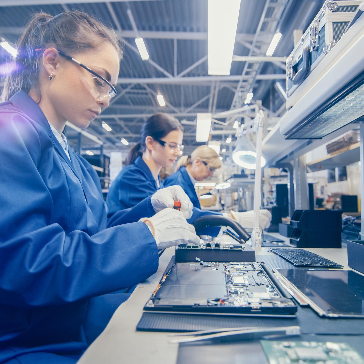 Woman Electronics Factory Worker in Blue Work Coat and Protective Glasses is Assembling Laptop's Motherboard with a Screwdriver. High Tech Factory Facility with Multiple Employees.