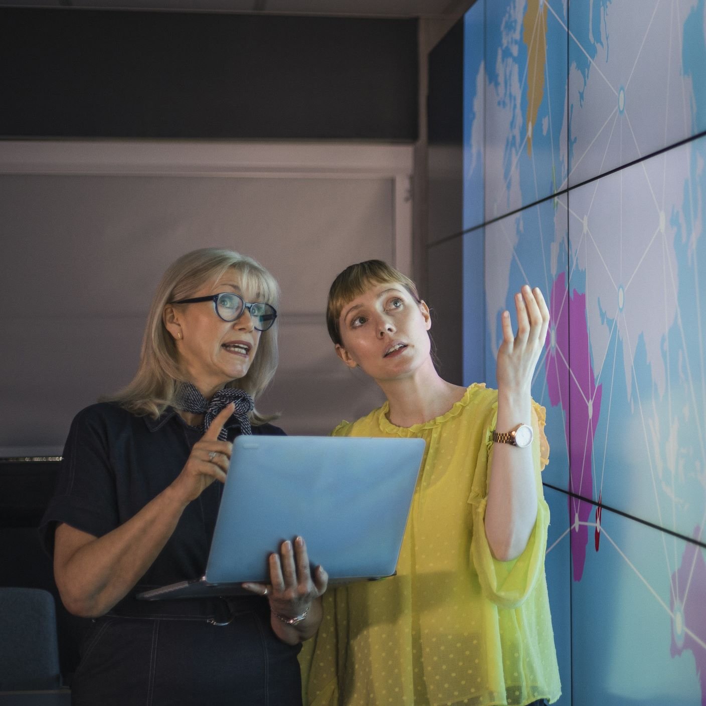 An experienced woman mentors a female colleague, the mature woman is holding a laptop as they debate data from an interactive display; they are both wearing smart casual clothing.