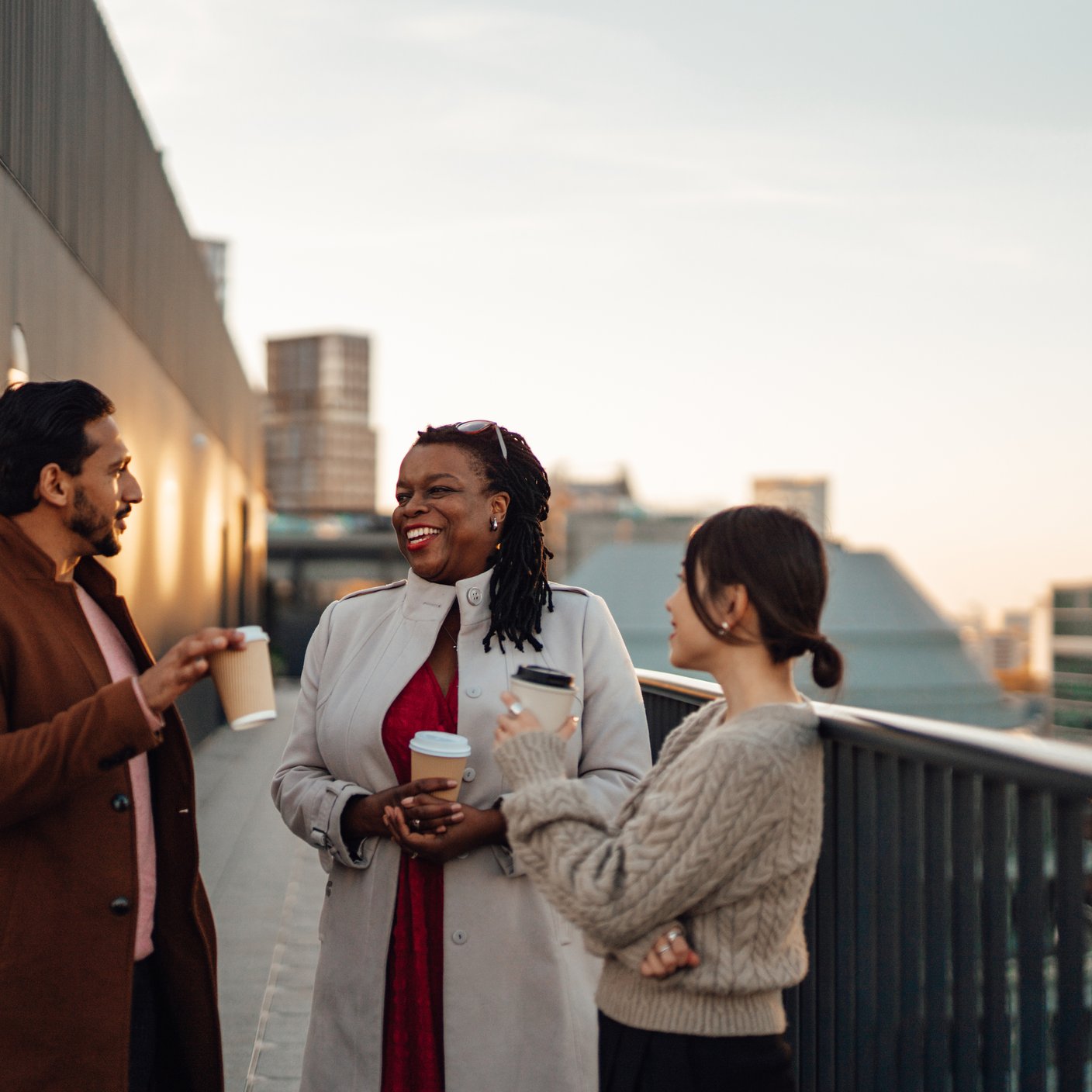 Cheerful multi-ethnic business people standing by railing at rooftop during coffee break. Talking about work and life together.
