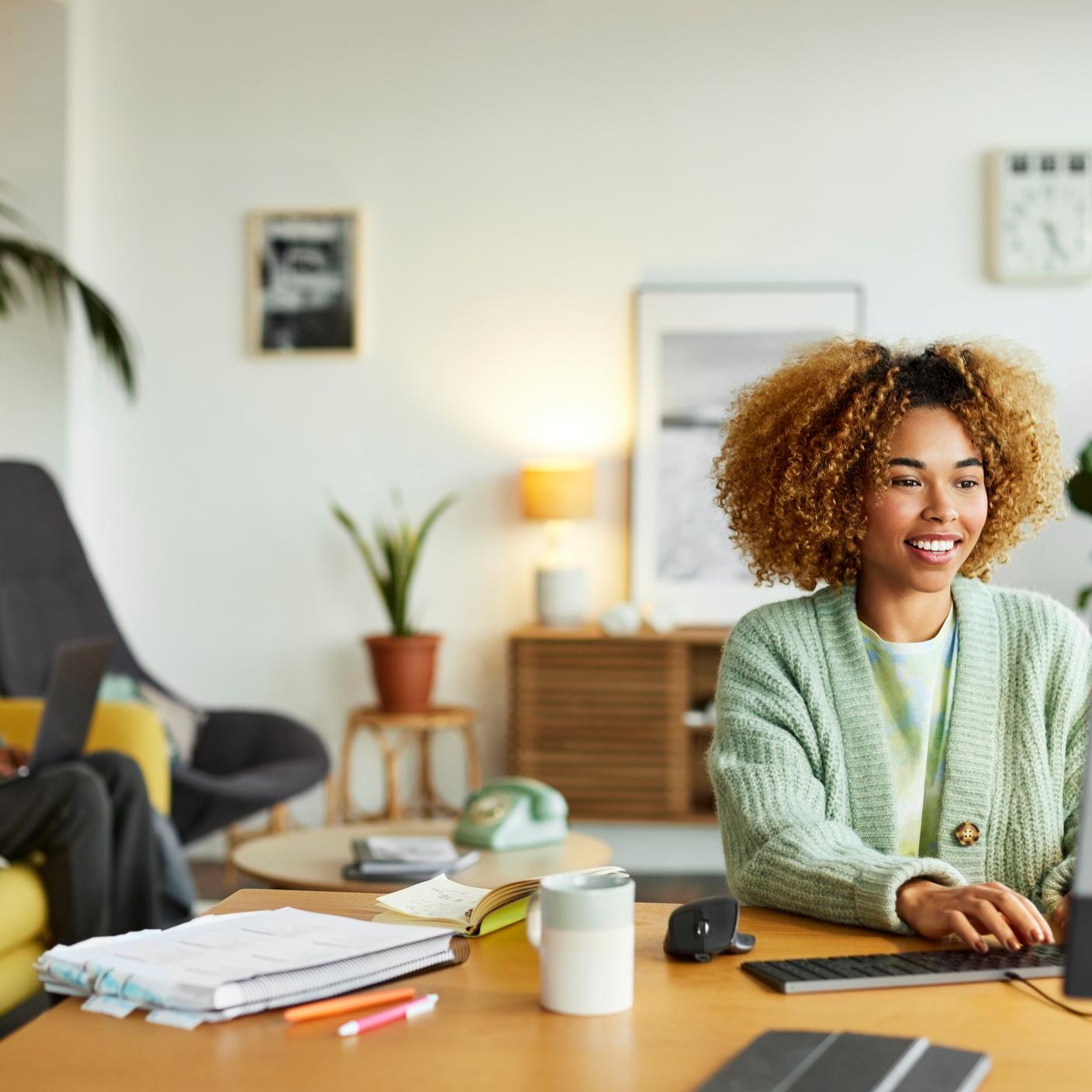 Smiling female freelancer using computer while colleague in background. Businesswoman is working at desk in home office. They are in apartment.