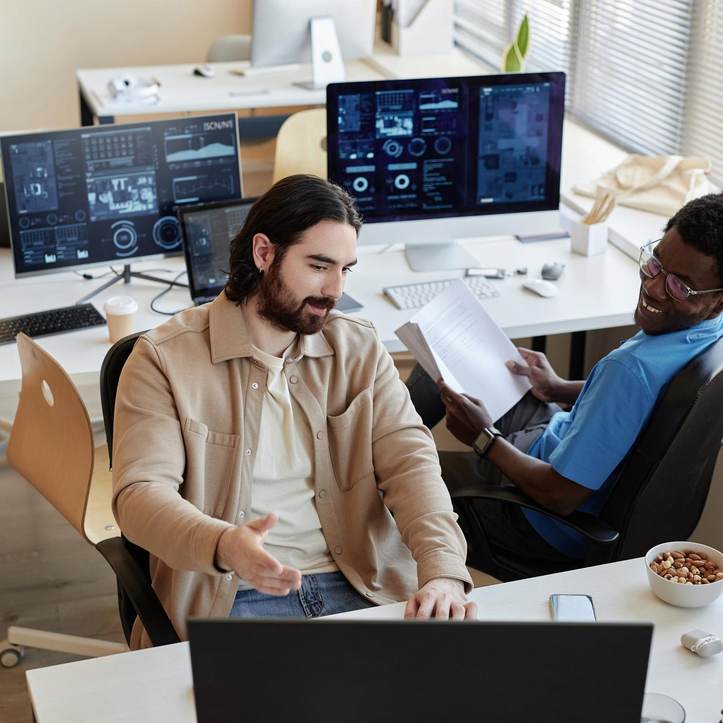 Young businessman showing data on computer screen to smiling African American colleague while explaining him results of analyzing it