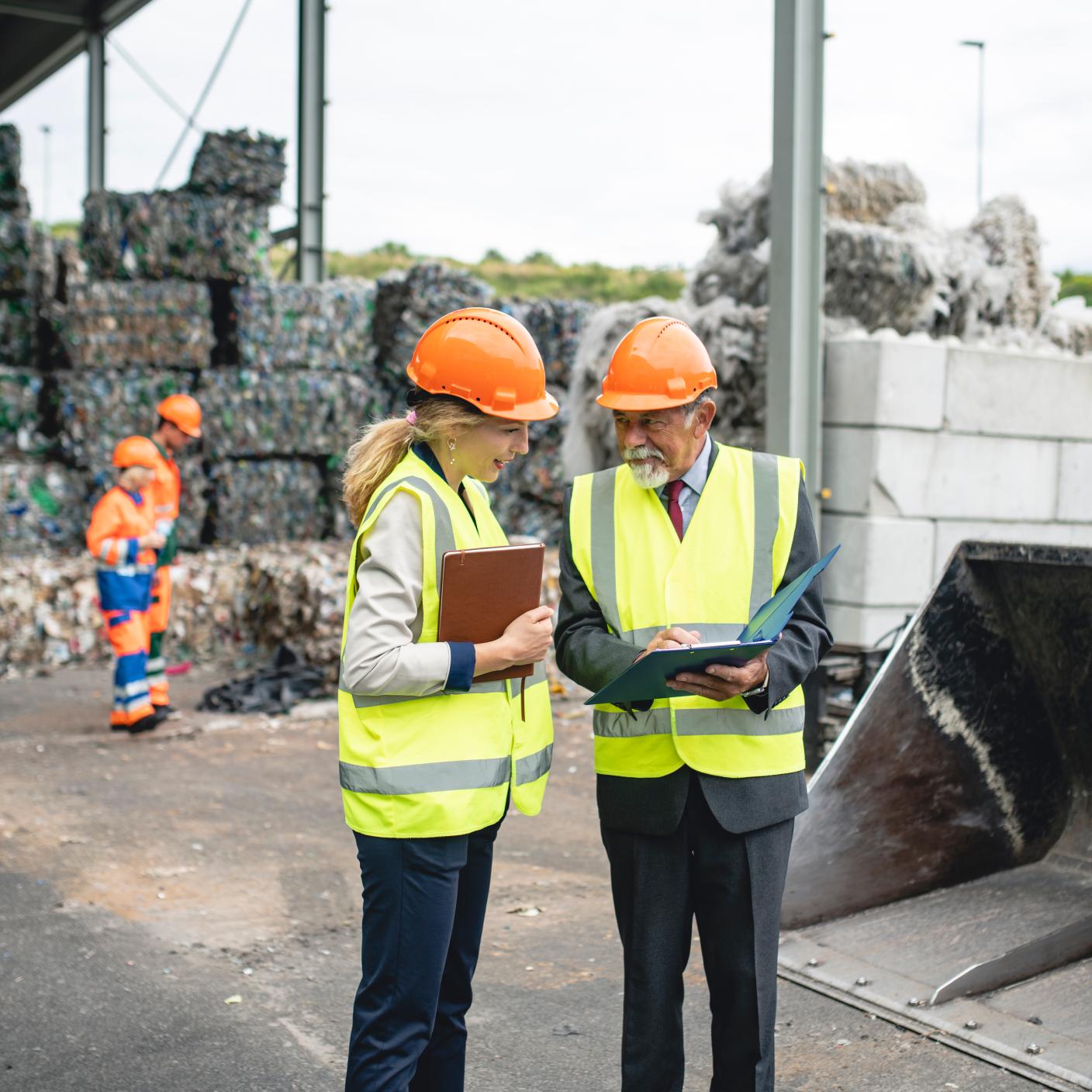 Quality control managers wearing reflective vests and helmets discussing findings outdoors at waste management facility