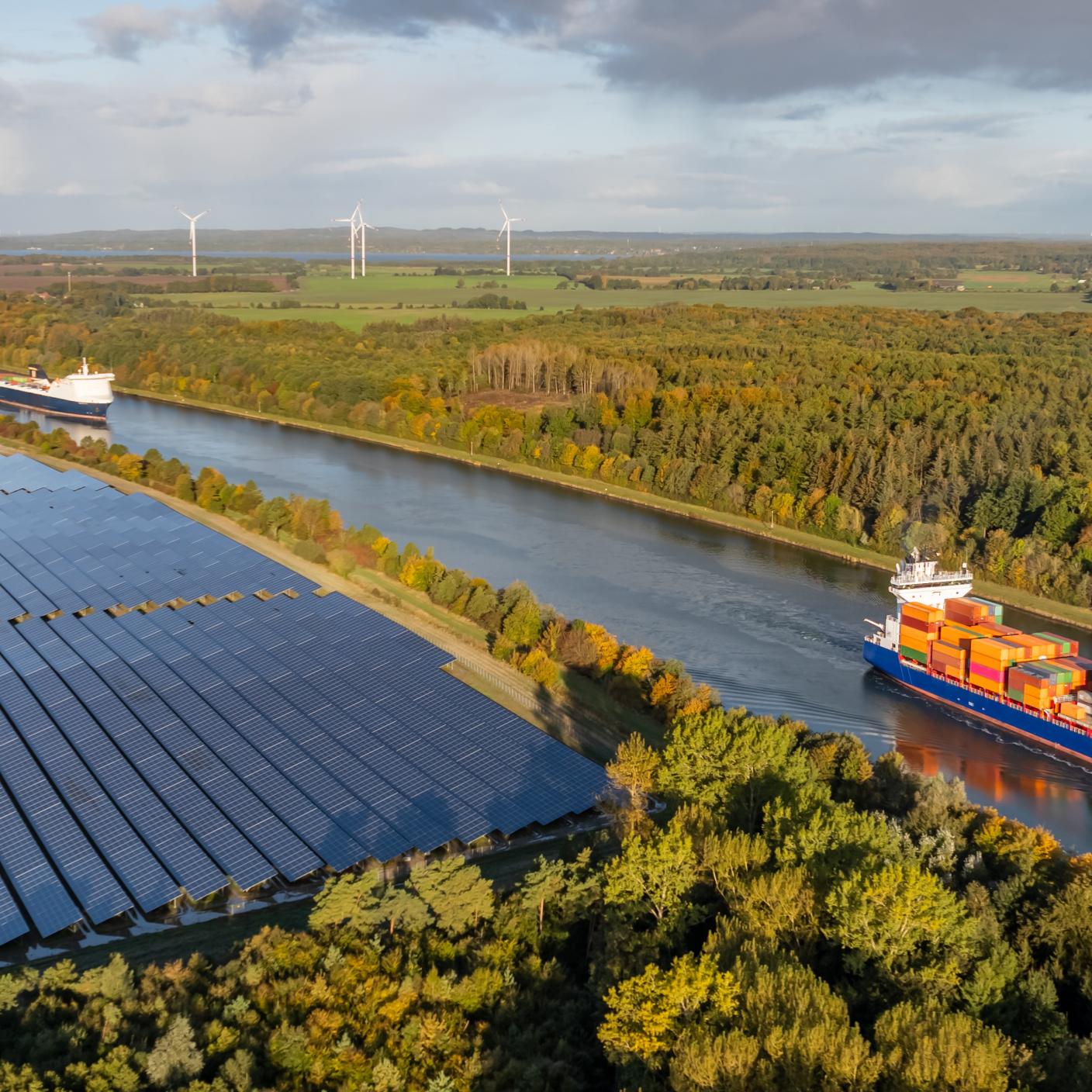 Container ship and car ferry navigate the Kiel Canal alongside vast solar panel fields and wind turbines, set against a backdrop of forest.