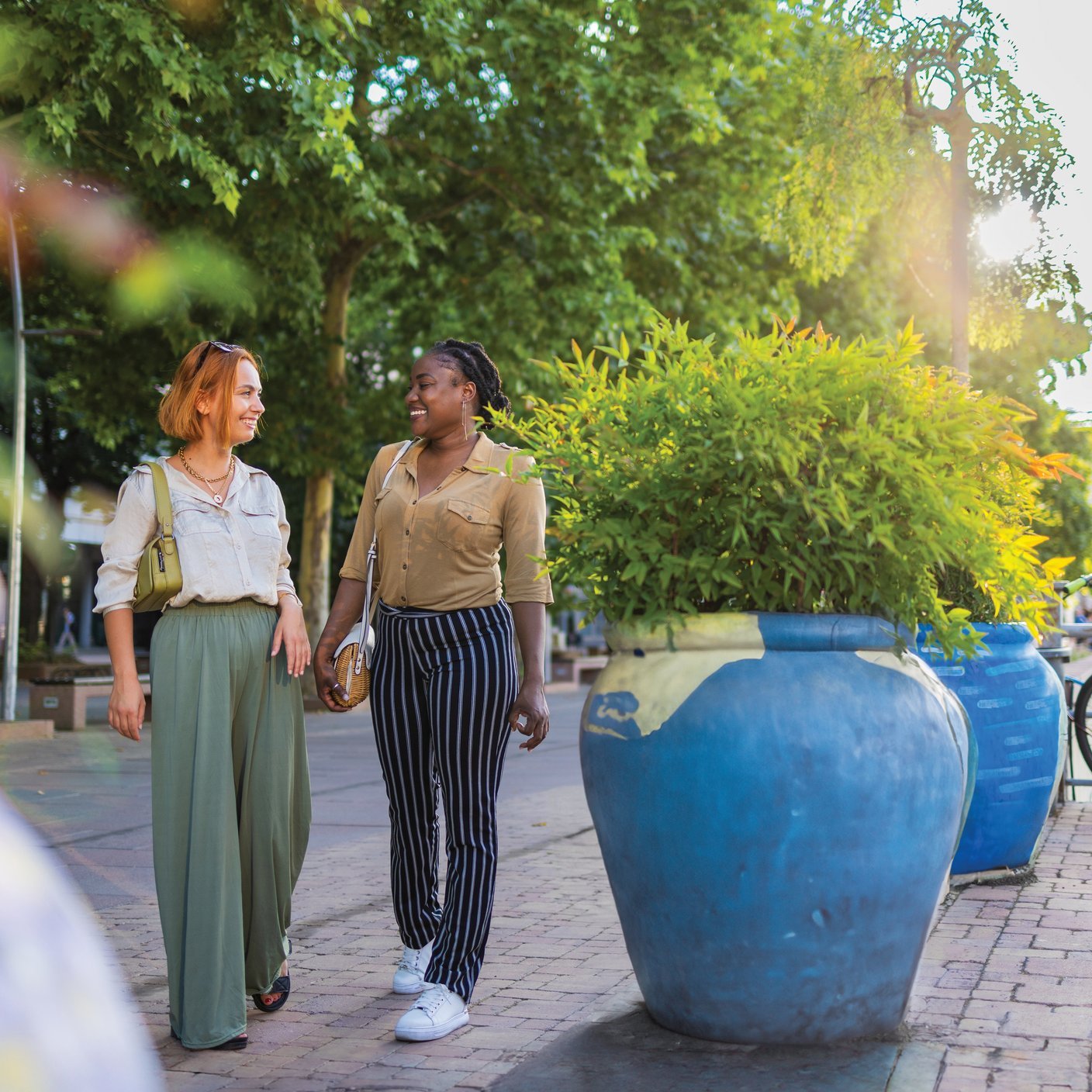 Two female friends walking having a conversation. They are located on an urban riverbank filled with greenery.