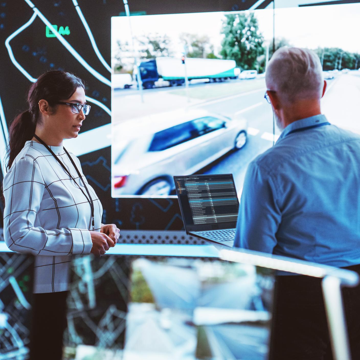 Modern Bir İzleme Ofisinde Büyük Bir Ekranın Yanında Konuşan Farklı Etnik Kökenli İki Yazılım Mühendisi ve Trafik Durumunu Gösteren Canlı CCTV Görüntüleri