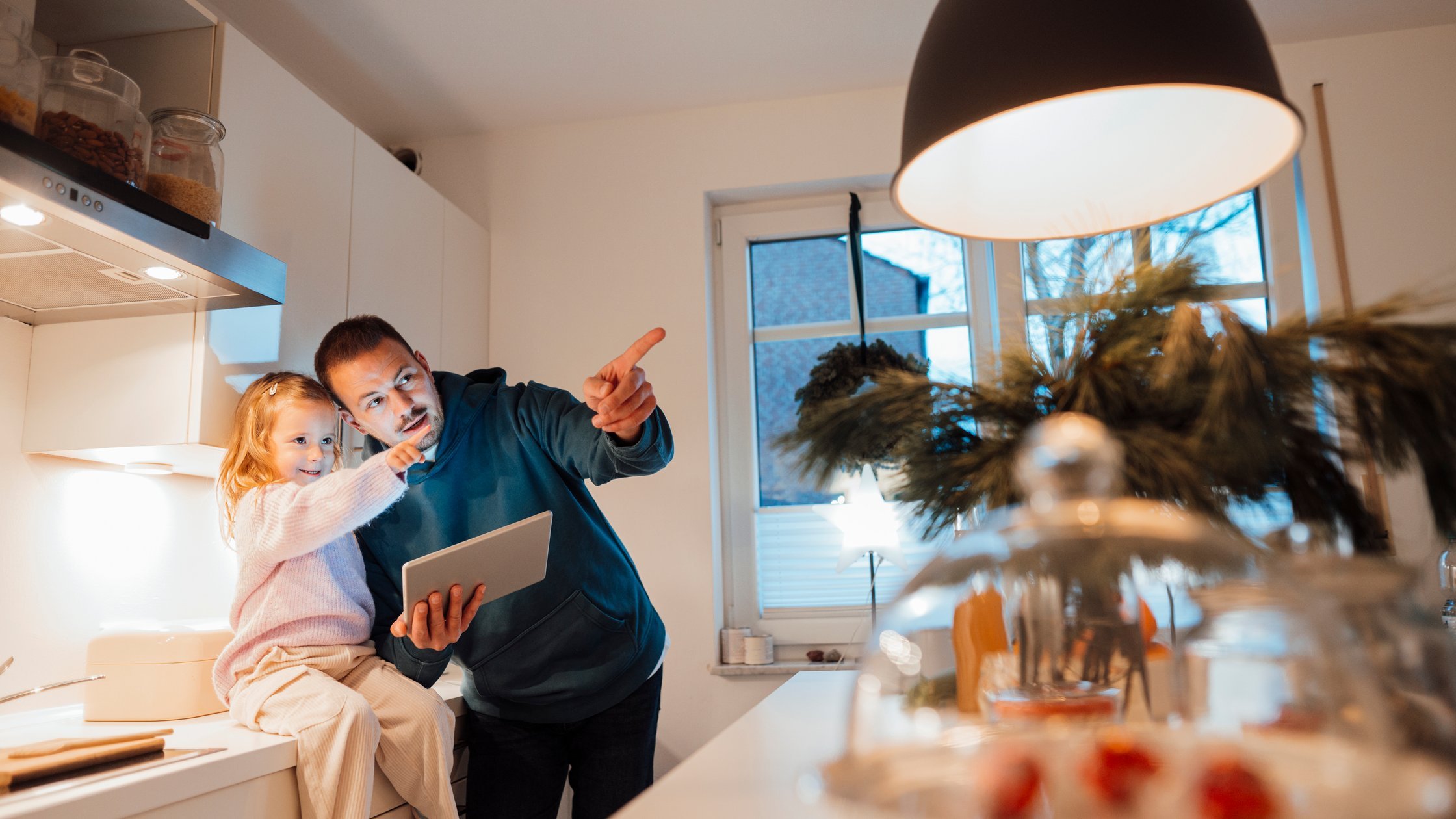 Daughter sitting on kitchen counter by father holding tablet PC gesturing at home.